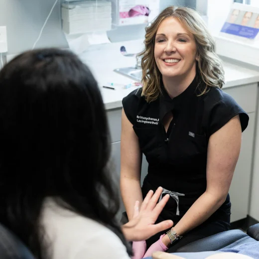 Smiling esthetician in black scrubs and pink gloves holding a hand mirror during a treatment-room consultation with a client
